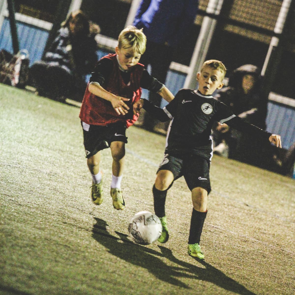 Two young players running with the ball across a floodlit pitch